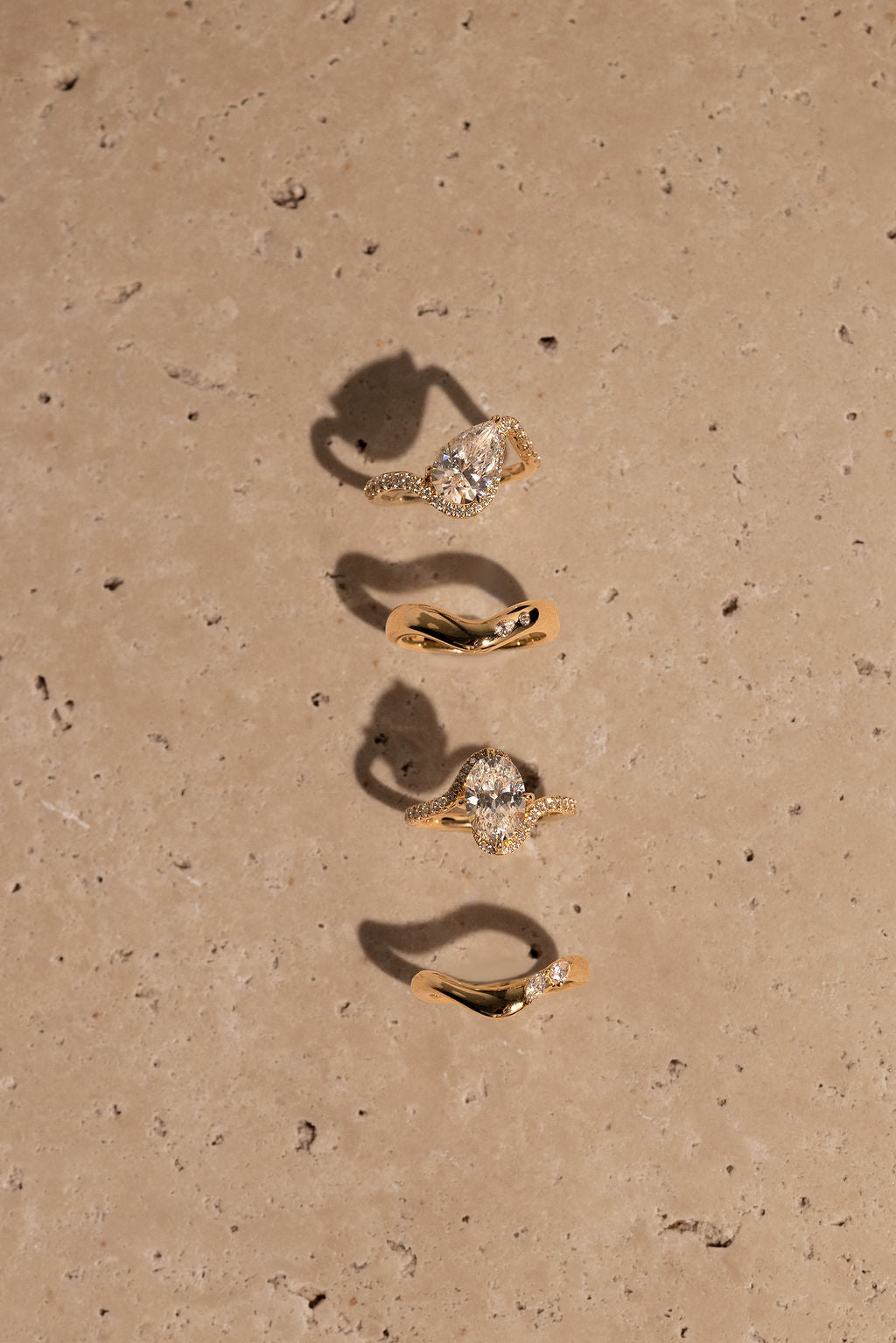 Four gold rings with diamond accents arranged vertically on a beige surface.