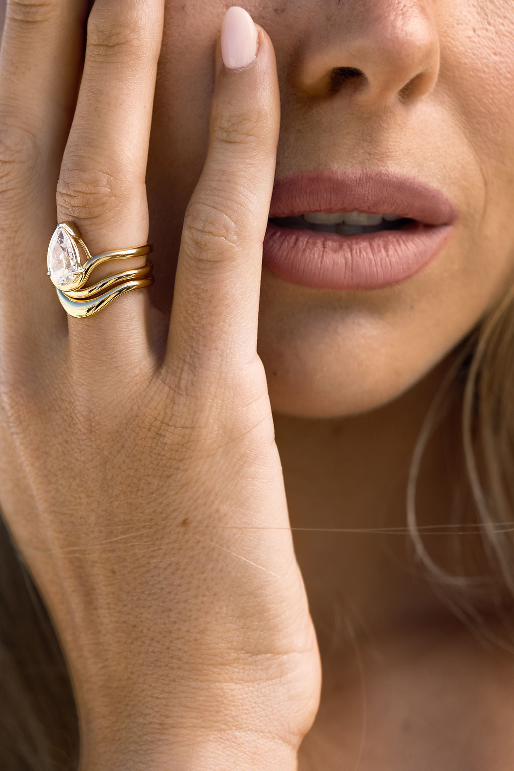 Close-up of a hand wearing a gold ring with a pear-shaped diamond, touching the face.