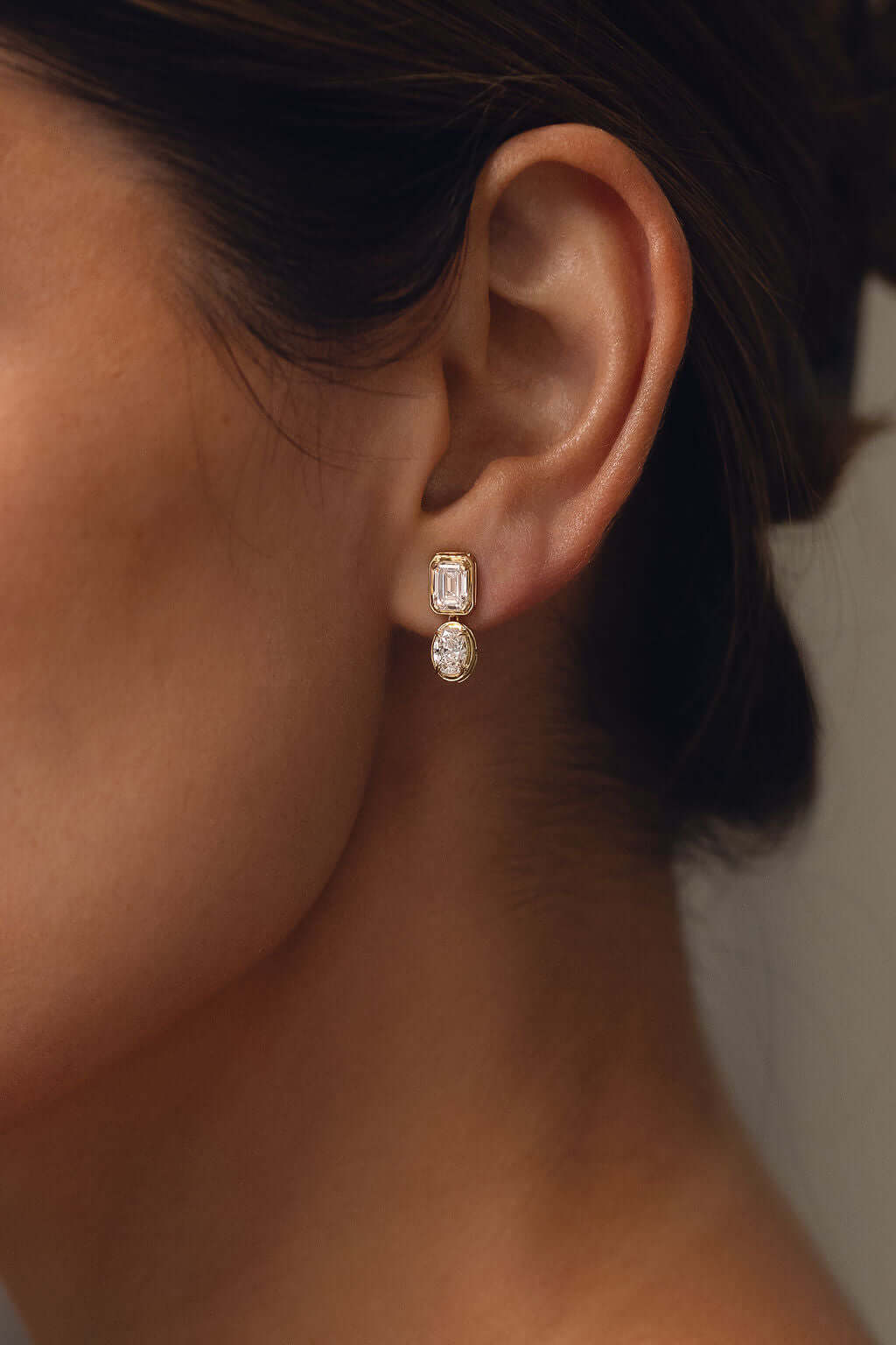 Emerald and Oval Bezel Stud Earrings Lab Diamond displayed on model's ear in yellow gold setting.