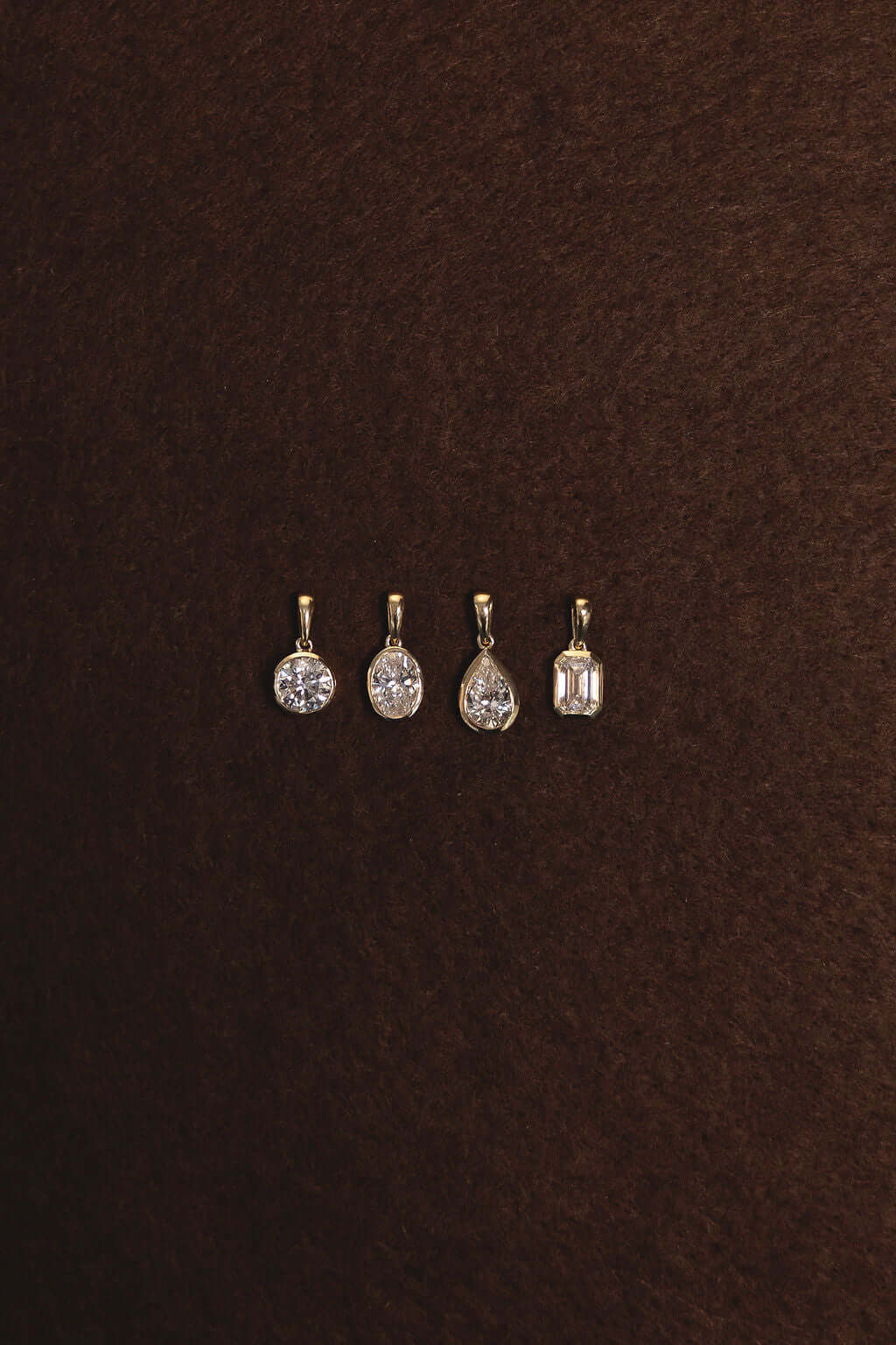 Four 1ct Oval Lab Diamond Bezel Pendants in yellow gold displayed on a brown background.
